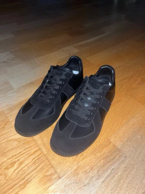Svarta sneakers från Maison Margiela - Säljer ett par svarta sneakers från Maison Margiela. Skorna har en stilren design med ovandel i mocka och läder, samt matchande svarta skosnören. Klassisk siluett som passar till många olika outfits.