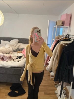 Gul omlottopp med knyt - Trendig gul omlottopp med knyt framtill och lång ärm. Toppen har en djup v-ringning och är i ett mjukt, stretchigt material som sitter snyggt på kroppen. Perfekt att styla med linne under för en cool lager-på-lager-look.