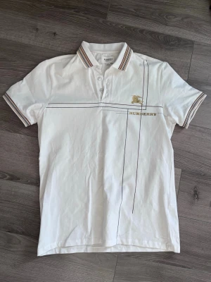 Vit pikétröja från Burberry - Vit pikétröja från Burberry med klassisk krage och knappar framtill. Tröjan har tunna linjer i svart och guld samt Burberry-logga på bröstet. Kragen och ärmsluten har randiga detaljer i beige, röd och svart. Perfekt för en stilren look.