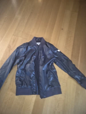 Mörkblå bomberjacka i glansigt tyg - Snygg mörkblå bomberjacka i glansigt material med ribbade muddar och krage. Jackan har dragkedja framtill, fickor med tryckknapp och en klassisk bomberpassform. Perfekt för dig som vill ha en stilren och trendig look.