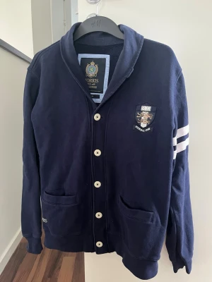 Marinblå cardigan från Morris - Snygg marinblå cardigan från Morris med vita knappar och två vita ränder på ena ärmen. Tröjan har två fickor framtill och ett broderat märke på bröstet. Skön krage och klassisk collegestil, perfekt för en avslappnad look.