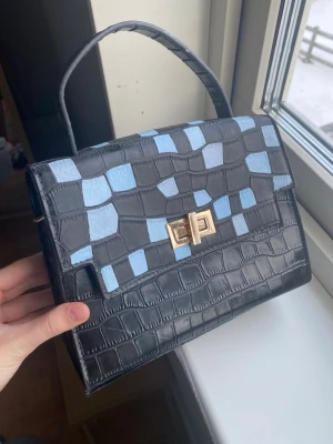 VINTAGE Svart handväska med blått mönster - Snygg svart handväska i krokopräglat konstläder med ljusblå rutmönstrade detaljer. Väskan har ett stilrent handtag, guldfärgat vridlås och metallfötter undertill. Perfekt accessoar för att lyfta din outfit.