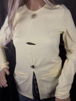 Ljusgul kofta med dekorativa knappar - Trendig ljusgul kofta med unika metallknappar och en snygg broschdetalj vid halsen. Koftan har rund halsringning, långa ärmar och två fickor framtill. Materialet är ribbat och ger en stilren look som passar perfekt till jeans.