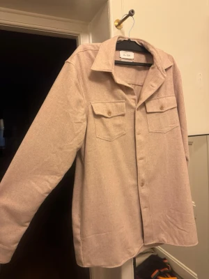 Beige overshirt - Stilren beige overshirt från Van Gils med klassisk krage och två bröstfickor med lock. Skjortjackan har raka ärmar och knäppning framtill. Materialet ser ut att vara mjukt och lite tjockare, perfekt för lager på lager. Enkel och clean design som passar till mycket.
