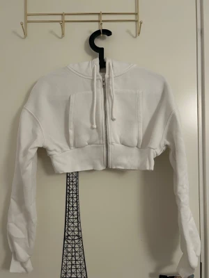 Vit croppad hoodie med dragkedja från Pull & Bear  - Vit croppad hoodie med dragkedja framtill och stora fickor från Pull & Bear i storlek S. Hoodien har ribbade muddar och justerbar huva med snören. OBSERVERA och se bilder på att hoodien är nopprig, därav priset. 