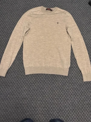 Beige stickad tröja från Morris  - Snygg beige stickad tröja från Gant med rund halsringning och diskret logga broderad på bröstet. Tröjan har långa ärmar och ribbade muddar vid ärmslut och nederkant. Perfekt till lager på lager och enkel att matcha.