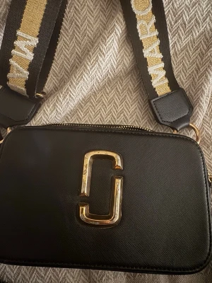 Svart axelväska från Marc Jacobs - Snygg svart axelväska från Marc Jacobs med guldiga detaljer och bred axelrem med logotyp. Väskan har en rektangulär form och är tillverkad i skinn med två dragkedjor upptill. Perfekt för att lyfta din outfit.