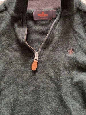 Grön stickad halvzip-tröja Morris - Mörkgrön stickad tröja från Morris i 100% merinoull. Halvzip med snygg dragkedja och läderdetalj, samt broderad logga på bröstet. Perfekt för lager-på-lager och har en klassisk, tidlös look.
