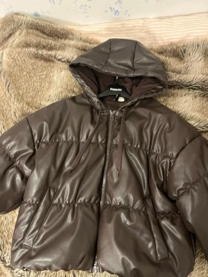 Brun pufferjacka från H&M - Snygg brun pufferjacka från H&M i glansigt material med huva och dragkedja framtill. Jackan har två sidofickor och dragsko i nederkant för extra passform. 