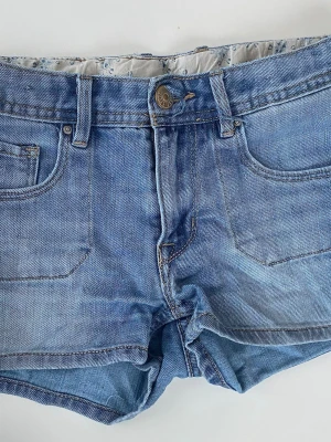 Blå jeansshorts med fem fickor - Snygga blå lågmidjade jeansshorts i klassisk modell med fem fickor och bälteshällor. Shortsen har knapp och dragkedja framtill samt en avslappnad passform. Perfekta till sommaren och lätta att matcha med olika stilar.