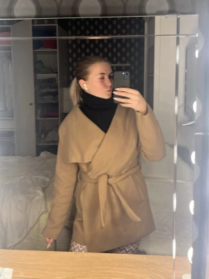 Beige omlottkappa med knytskärp - Stilren beige kappa i omlottmodell med brett slag och knytskärp i midjan. Kappan har en rak passform och är perfekt för lager-på-lager. Materialet ser ut att vara mjukt och lite tjockare, vilket ger en lyxig känsla.