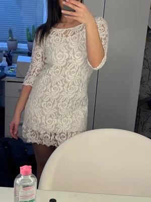 Vit figurnära miniklänning i spets  - Söt, vit miniklänning med vita spetsrosor. Klänningen har en vit underklänning vilket gör att den inte är genomskinlig. Perfekt som studentklänning eller inför annat tillfälle.
