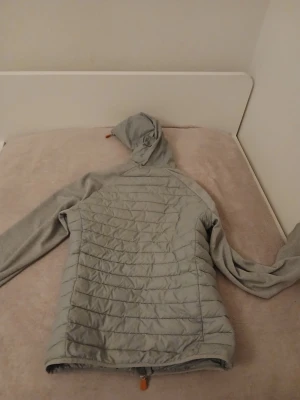 Ljusgrå Save the duck jacka med huva - Ljusgrå vindjacka med quiltad front och rygg, samt mjuka ärmar i sweatshirtmaterial. Jackan har hel dragkedja framtill, två sidofickor och en justerbar huva. Perfekt för lager-på-lager och chill stil.