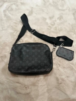 Riktigt snygg  svart axelväska  - Snygg svart axelväska från med klassiskt monogrammönster i grått. Väskan har justerbar axelrem med logga och en liten avtagbar miniväska. Tillverkad i tåligt läder och perfekt för dig som vill ha en trendig och praktisk accessoar.