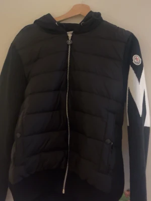 Svart cardigan med huva från Moncler - Snygg svart cardigan från Moncler med huva och dragkedja framtill. Jackan har stickade ärmar med vit M på ärmen och Moncler-logga på ena ärmen. Perfekt för kalla vinterdagar och riktigt trendig look. Pris kan diskuteras 