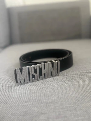 Svart Moschino bälte med silverdetalj - Snyggt svart bälte från Moschino med stor silverfärgad logotyp som spänne. Bältet är stilrent och tillverkat i skinn, perfekt för att lyfta vilken outfit som helst med en statement-detalj. De är lite repor på lågan men de ser anda bra ut. De är typ 110 cm tror jag.