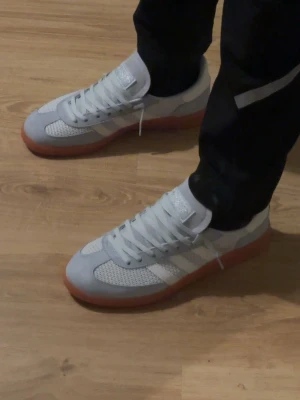 Adidas handball spezials  - Snygga Adidas sneakers i vitt och ljusgrått med klassiska tre ränder på sidan. Ovandelen är i mesh och mocka för en fräsch look, och sulan är i gummi med brun ton. Perfekta för dig som gillar stilrena och sportiga skor.