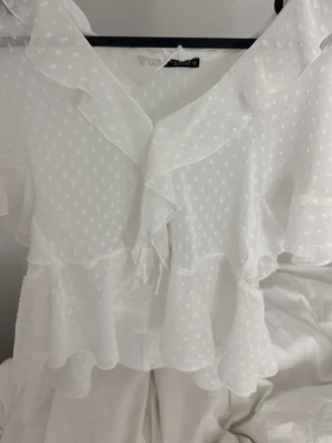 Vit volangblus från Zara - Säljer en söt vit blus från Zara med volanger och prickigt strukturmönster. Blusen har v-ringning och korta ärmar med volangdetaljer. Tunt, lätt och transparent material som ger en romantisk känsla. Perfekt att styla med jeans eller kjol.
