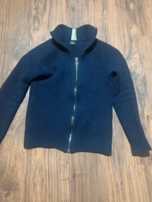 Mörkblå stickad ziptröja från BOSS - Mörkblå stickad tröja från BOSS med hel dragkedja framtill och hög krage. Tröjan har ett diskret rutmönster i stickningen och ribbade muddar vid ärmslut och nederkant. Perfekt för lager-på-lager och snygg till jeans.