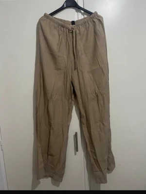 Beiga linnebyxor med dragsko - Snygga och luftiga beiga linnebyxor med resår och dragsko i midjan. Perfekta för en avslappnad stil och sköna att bära under varma dagar. Byxorna har raka ben och är tillverkade i ett lätt linnematerial.