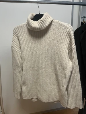 Beige ribbstickad polotröja - Mysig beige ribbstickad polotröja med bred krage och avslappnad passform. Tröjan har långa ärmar och är tillverkad i ett mjukt, tjockt material som passar perfekt för kyliga dagar. Enkel och stilren design utan synligt märke.