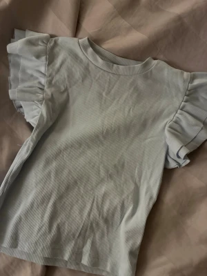 Ljusblå ribbad topp från Gina Tricot - Supersöt ljusblå topp från Gina Tricot med ribbad struktur och volangärmar. Perfekt som en enkel men ändå trendig look. Toppen har rund halsringning och är bara använd ett fåtal gånger