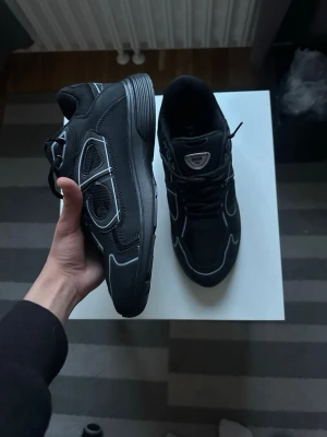 Dior b30 - Säljer ett par svarta sneakers från Dior med coola silvriga detaljer och meshpaneler. Skorna har en chunky sula och klassisk snörning. Perfekta för dig som vill ha en stilren men ändå sportig look.