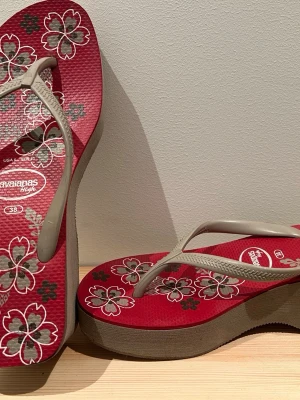 Röda Havaianas platåsandaler blommiga - Snygga röda platåsandaler från Havaianas med beige remmar och blommigt mönster i vitt och grått på sulan. Tillverkade i gummi för skön komfort och cool look. Perfekta för dig som vill ha lite extra höjd och en färgglad touch till din outfit.