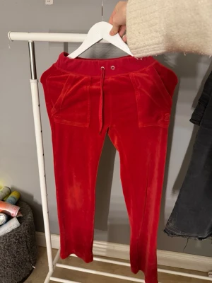 Röda mjukisbyxor från juicy couture - Röda mjukisbyxor i mjuk velour med snörning i midjan och stora fickor fram. Bakfickor med lock och tryckknappar. Perfekta för en chill och avslappnad stil.