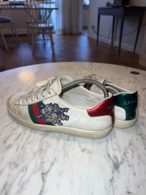 Gucci Ace sneakers med brodyr - Gucci Ace sneakers i vitt skinn med klassiska gröna och röda ränder på sidan samt blå broderad detalj. Baksidan har metallicröd och metallicgrön häl med Gucci-logga. Skorna har snörning och platt sula.