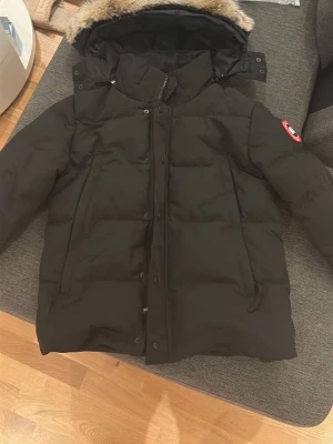 Canada Goose Wyndham - Säljer en svart dunjacka från Canada Goose med huva och fuskpäls. Jackan har klassisk pufferstil, dragkedja och knappar framtill samt logga på ärmen. Perfekt för kalla vinterdagar och riktigt snygg streetstil.