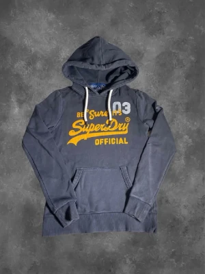 Mörkgrå Superdry hoodie med tryck - Mörkgrå hoodie från Superdry med stor gul och vit logga på bröstet och texten 'Be Sure It's SuperDry Official'. Hoodien har vita dragsnören, känguruficka och ribbade muddar. Perfekt för en avslappnad streetwear-look.