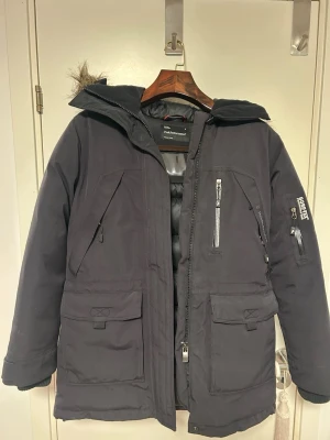 Svart Expedition parkas från Peak Performance - Säljer en svart Expedition parkas från Peak Performance med GORE-TEX Performance Shell. Jackan har flera fickor med dragkedja, justerbar huva med avtagbar fuskpäls och robust dragkedja framtill. Perfekt för kalla dagar. Rök och djurfritt. Sparsamt använd. Ord.pris 6500
