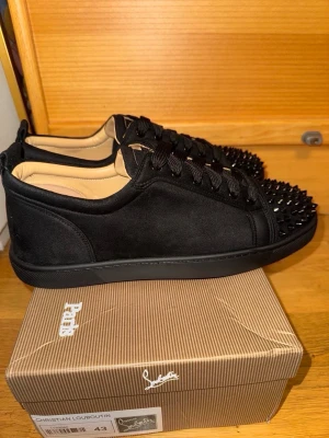 Svarta Christian Louboutin sneakers - Snygga svarta sneakers från Christian Louboutin med mattsvarta nitar på tån och den ikoniska röda sulan. Skorna är låga och har en ovandel i mocka med svarta skosnören och diskreta detaljer. Perfekta för dig som vill sticka ut med exklusiv stil.