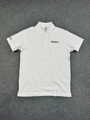 Vit piké från Stadium med tryck - Klassisk vit pikétröja från Stadium med korta ärmar och krage. Tröjan har knappar framtill och Stadium-logga tryckt på bröstet och ena ärmen. Perfekt till en clean och sportig look.