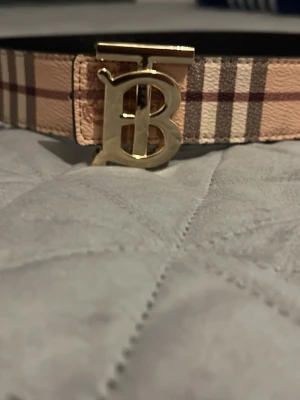 Burberry bälte med guldigt B-spänne - Snyggt Burberry bälte med klassiskt rutigt mönster på ena sidan och svart på andra. Bältet har ett stort guldigt B-spänne som ger en lyxig känsla. Perfekt accessoar för att lyfta din outfit.