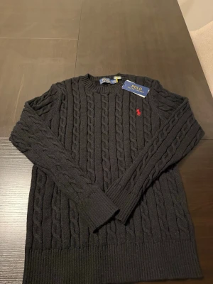 Svart kabelstickad tröja Polo Ralph Lauren - Kabelstickad svart tröja från Polo Ralph Lauren med rund halsringning och röd broderad logga på bröstet. Tröjan har långa ärmar och ribbade muddar vid ärmslut och nederkant. Perfekt för dig som gillar klassisk och stilren design.