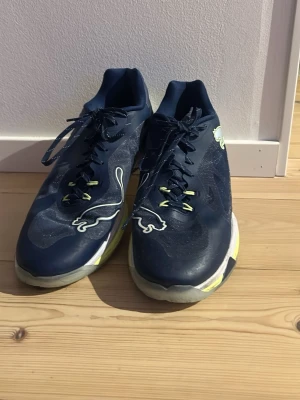 Marinblå Puma inomhussko med neon - Snygga marinblå träningsskor från Puma med stora vita och neongula loggor på sidorna. Skorna har blåa snören, meshdetaljer och en vit sula med inslag av neon. Perfekta för dig som tränar inomhus! Använda i 3 veckor ungefär.