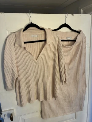 Beige ribbstickat set från Stockh LM - Säljer ett beige ribbstickat set från Stockh LM i storlek M. Setet består av en kortärmad tröja med krage och v-ringning samt en matchande kjol. Båda plaggen har en mjuk och stretchig känsla, perfekta för en chill och stilren look.