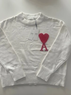Vit Ami sweatshirt S - Vit stickad tröja från Ami med ribbad rund hals och muddar. På bröstet finns ett stort rött hjärta och bokstaven A i ett coolt mönster. Riktigt stilig nu till vinter 
