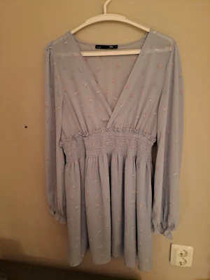 Ljusblå blommig omlottklänning - Ljusblå kortklänning med små blommor, omlott framtill och smock i midjan. Klänningen har långa ärmar med resår vid ärmslut och en luftig kjol. 