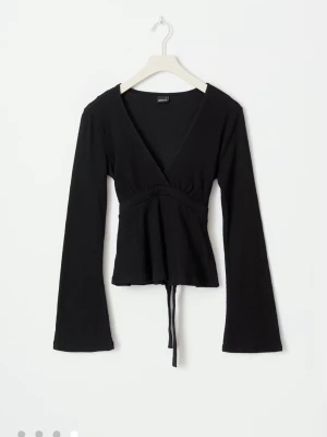 Svart omlottopp med knytband - Snygg svart omlottopp med v-ringning och långa ärmar. Toppen har knytband i midjan som ger en smickrande passform. Perfekt att styla med jeans eller kjol för en trendig look.