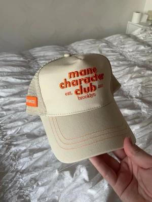 Beige truckerkeps från Amika - Snygg beige keps från Amika med orange brodyrtext 'mane character club' framtill och mesh på sidorna. Kepsen har böjd skärm, justerbar baksida och coola orange detaljer. 