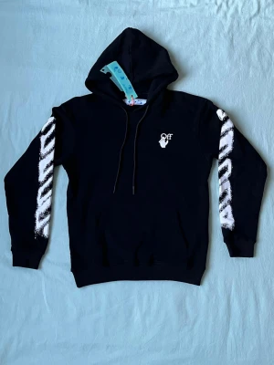 Svart Off-White hoodie med tryck - Svart hoodie från Off-White med vit logga och handmotiv på bröstet. Ärmarna har Off-Whites ikoniska vita diagonalränder med en borstad effekt. Klassisk huva med dragsko och känguruficka framtill. Perfekt för en streetwear-look.