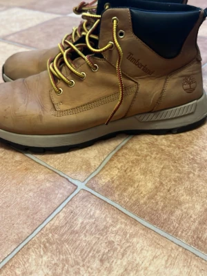 Bruna Timberland kängor i skinn - Säljer ett par snygga bruna Timberland kängor i skinn med gul- och rödmönstrade snören och svart vadderad kant upptill. Grov sula för bra grepp och klassisk Timberland-logga på sidan. Perfekta för dig som vill ha både stil och funktion.
