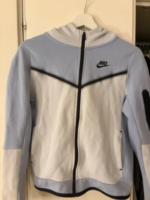 Nike ljusblå och vit hoodie - Nike hoodie i ljusblått och vitt med svart dragkedja och logga på bröstet. Hoodien har huva, svarta detaljer och en ficka med dragkedja på ärmen. Tillverkad i mjukt material, perfekt för en chill och sportig look. Storlek L i barnstorlek 