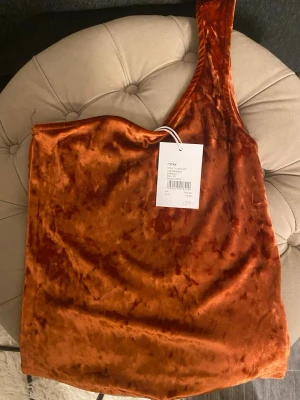 Rostorange oneshoulder sammetstopp - Säljer en snygg rostorange topp i mjuk sammet från Stradivarius. Toppen har en unik oneshoulder-design och är ärmlös, vilket ger en trendig och modern look. Perfekt för dig som vill sticka ut med en cool färg och skön känsla.