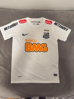 Neymar Jr vit santos fotbollströja - Säljer en vit Santos FC matchtröja från Nike med tryckta sponsorer i svart, orange och rött. Tröjan har klubbmärke på bröstet, Dri-FIT-material och korta ärmar. Perfekt för dig som älskar brasiliansk fotboll och vill ha en snygg supportertröja. PRIS KAN DISKUTERAS