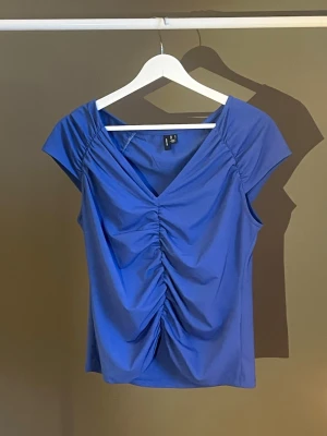Blå v-ringad topp med rynk framtill - Snygg blå topp med v-ringning och rynkdetalj framtill. Toppen har korta ärmar och är tillverkad i ett mjukt, stretchigt material som sitter bekvämt. Perfekt att styla med jeans eller kjol för en trendig look. Köpt från Vero Moda, använt ett par gånger💙