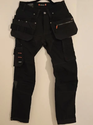 Svarta cargobyxor L.brador herr - Säljer ett par svarta cargobyxor från L.Brador med flera praktiska fickor, förstärkta knän och detaljer i grått. Byxorna har knappjustering vid benslut och robust design, perfekta för dig som gillar funktionella och slitstarka plagg.